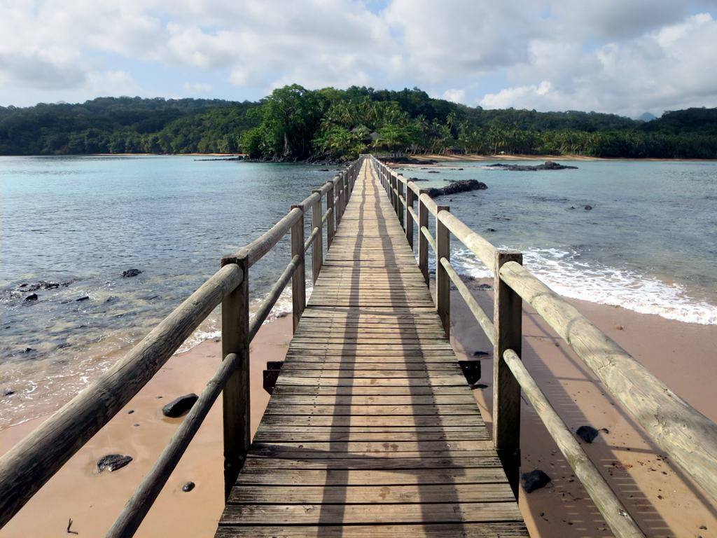 A wooden footbridge across clear water to the forested Bom Bom islet, Príncipe